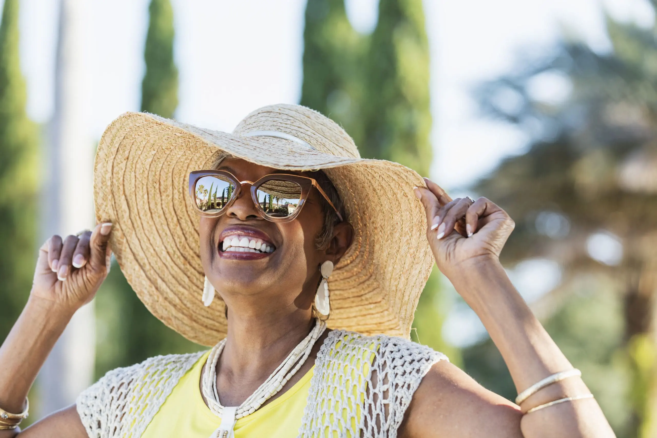 A beautiful senior African-American woman in her 70s wearing sunglasses and a wide brim hat on a sunny day. She is looking up at the sky, smiling. A building, palm trees and clear blue skies are visible in the reflection in the sunglasses.