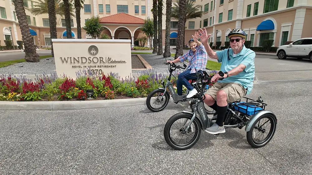 Two Windsor at Celebration residents ride electric adult tricycles past the community entrance sign, waving cheerfully as they stay active with fun mobility exercises for seniors.